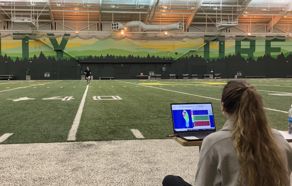 A person running on an athletic field while a woman (back facing camera) monitors the athlete on a computer