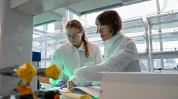Two people wearing white coats and safety goggles in a lab