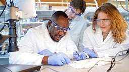 Three scientists in lab coats and protective earwear peer at a sample