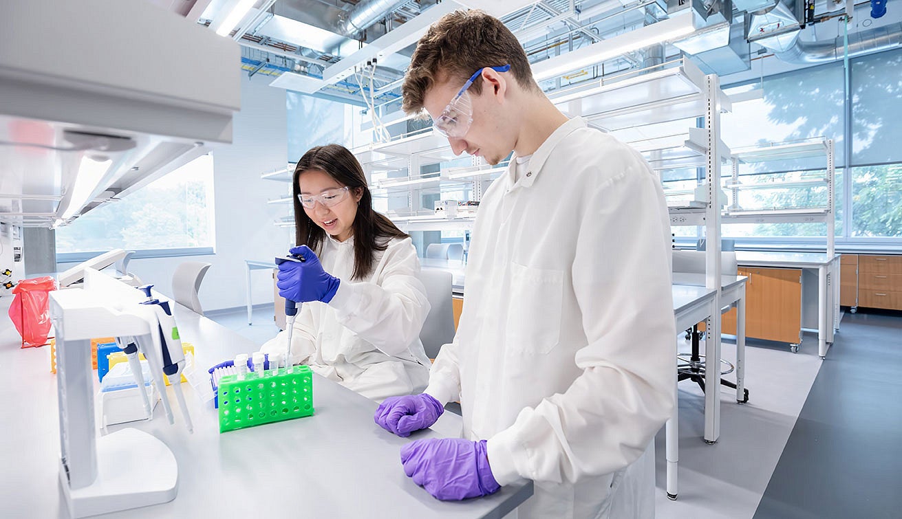 Young woman in lab coat pipetting while young man in lab coat looks on