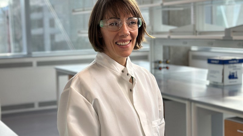 Woman in lab coat in lab with lab safety glasses smiling
